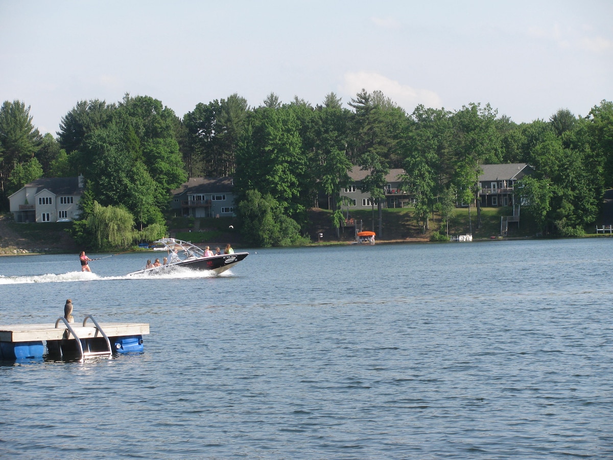 A scenic view of Little Sand Lake features a boat gliding across the water, with two individuals enjoying the ride. Lush green trees line the shoreline, where several residential structures are visible in the background. A dock extends into the lake, adding to the tranquil setting.