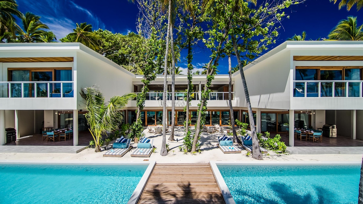 The villa features a modern architectural design, framed by lush palm trees and surrounded by tropical foliage. A spacious terrace with sun loungers extends towards a turquoise swimming pool, leading to a private beach area. Large glass windows provide a bright, open atmosphere.