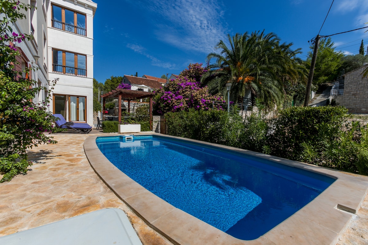 A private pool area is surrounded by lush greenery and vibrant flowers. The clear water of the pool reflects the blue sky above, while sun loungers are positioned alongside. A wooden structure with decorative foliage provides shade, enhancing the outdoor relaxation space.