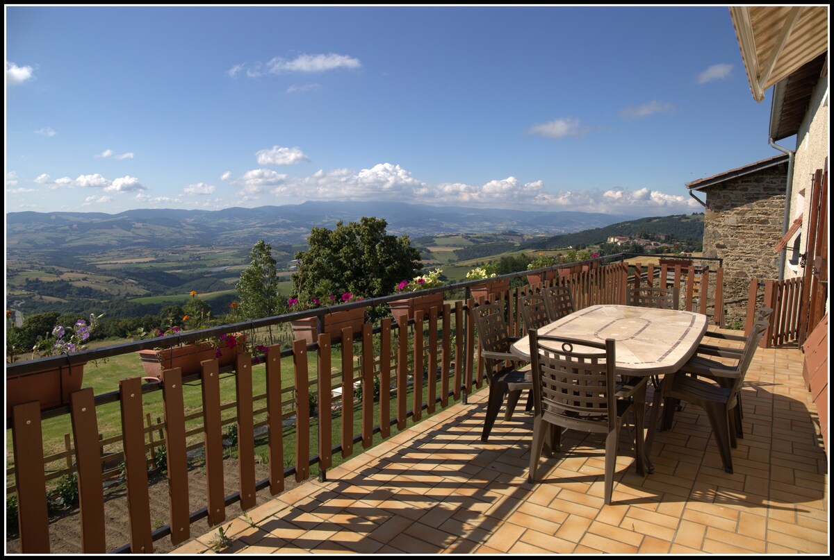 A spacious outdoor balcony features a large dining table surrounded by multiple chairs, offering expansive views of rolling hills and distant mountains. Decorative flowers add color to the railing, while sunlight casts playful shadows across the tiled floor.
