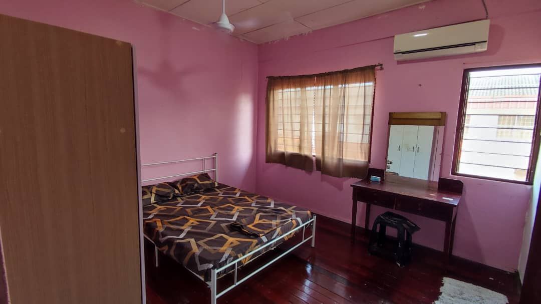A cozy bedroom features pink walls and dark wooden flooring. A bed with a geometric patterned comforter is positioned against one wall. Natural light enters through a window, complemented by curtains. A small desk and chair are situated near the window, alongside a freestanding wardrobe.