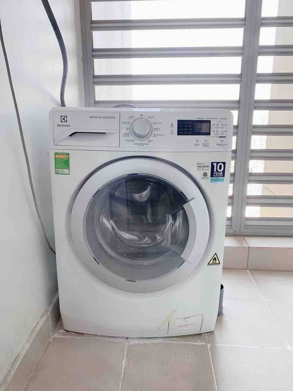 A modern front-loading washing machine is positioned near a window with horizontal blinds. The machine is white with a digital display and various control options, reflecting contemporary design. Natural light filters in, enhancing the clarity of the appliance's sleek surface.