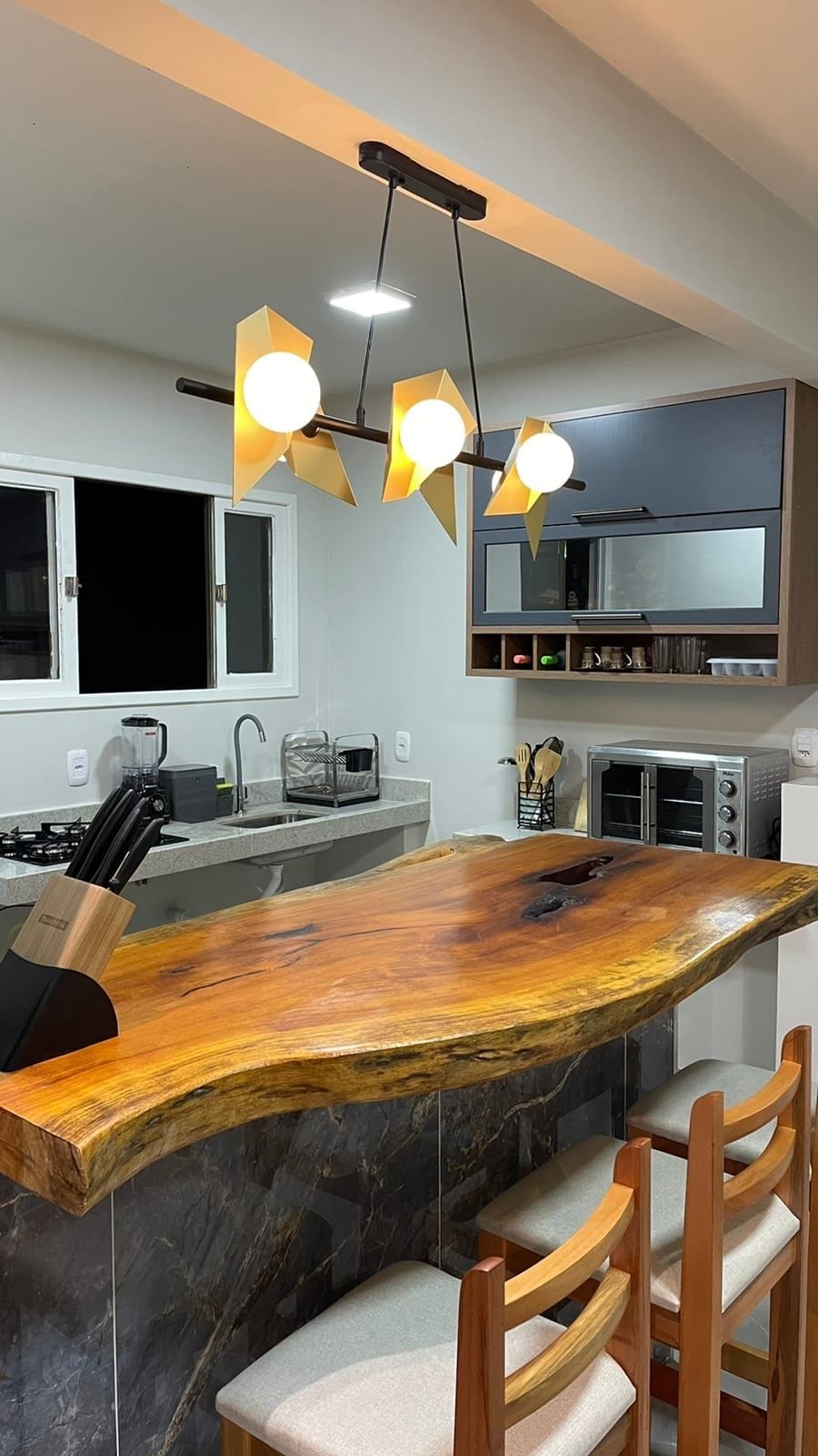 A modern kitchen is featured with a unique wooden countertop, providing a rustic touch. The area is illuminated by a stylish light fixture with three bulbs. A gas stove and sink are visible, along with wood cabinetry and several kitchen utensils.