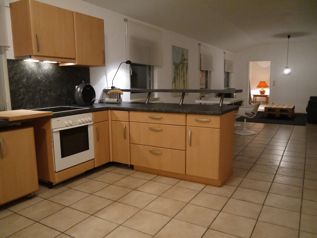 An open kitchen area features light wooden cabinetry and a dark countertop. A stovetop and oven are integrated into the cabinetry. The space is illuminated by overhead lighting, enhancing the tiled floor's neutral color. An opening leads into the living area where a lamp provides additional light.