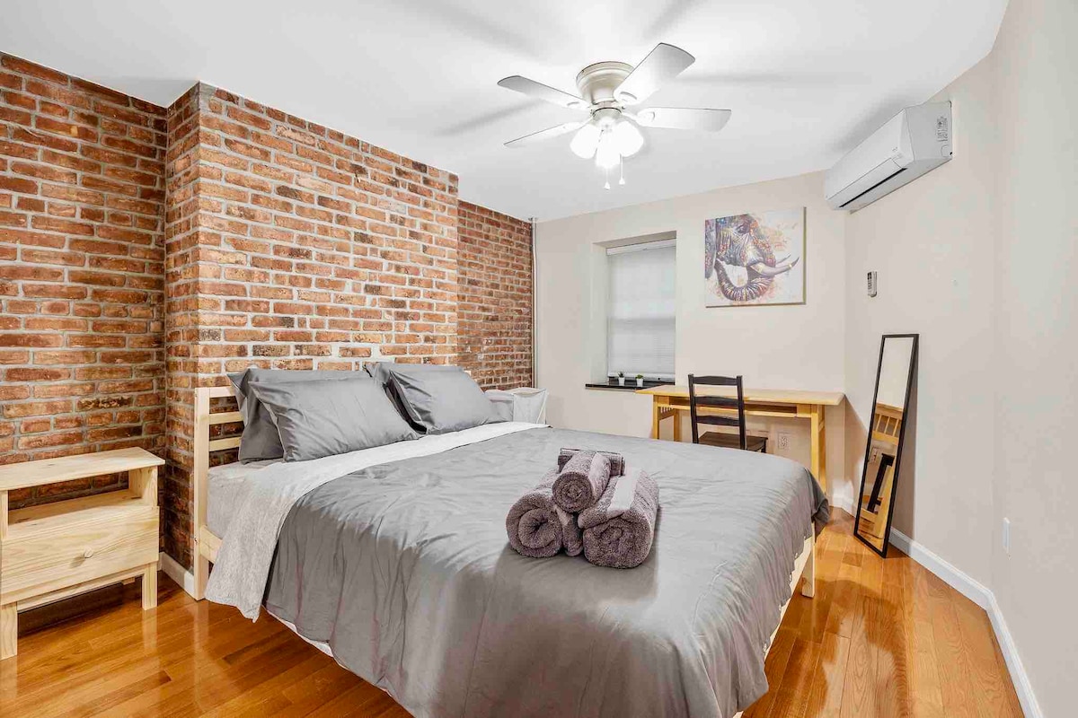 A spacious bedroom features exposed brick walls that provide a charming backdrop. A sizable bed is centered in the room, accompanied by a nightstand. A small desk and chair are situated beside a window, while a mirror reflects the inviting atmosphere.