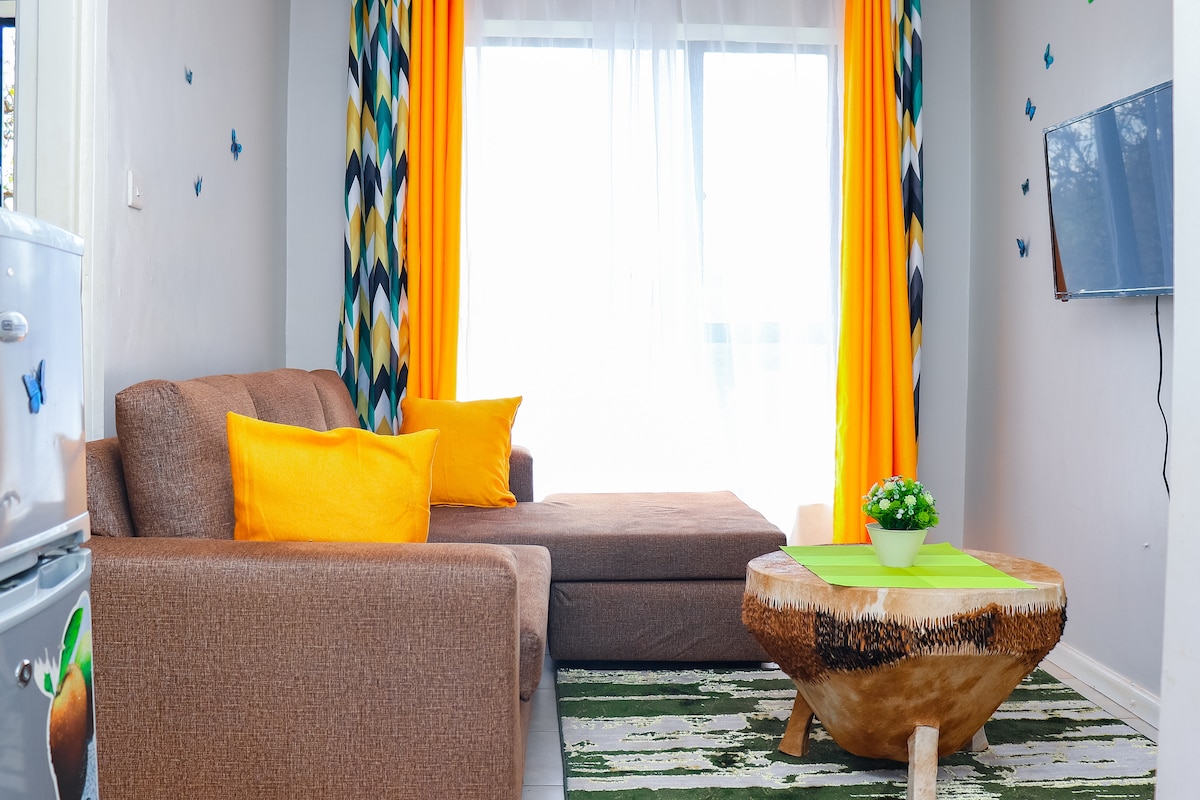 A cozy living area is furnished with a comfortable brown couch adorned with bright yellow pillows. A round wooden table sits in front, complemented by a green table runner. Large windows draped with vibrant, patterned curtains allow natural light to brighten the space.