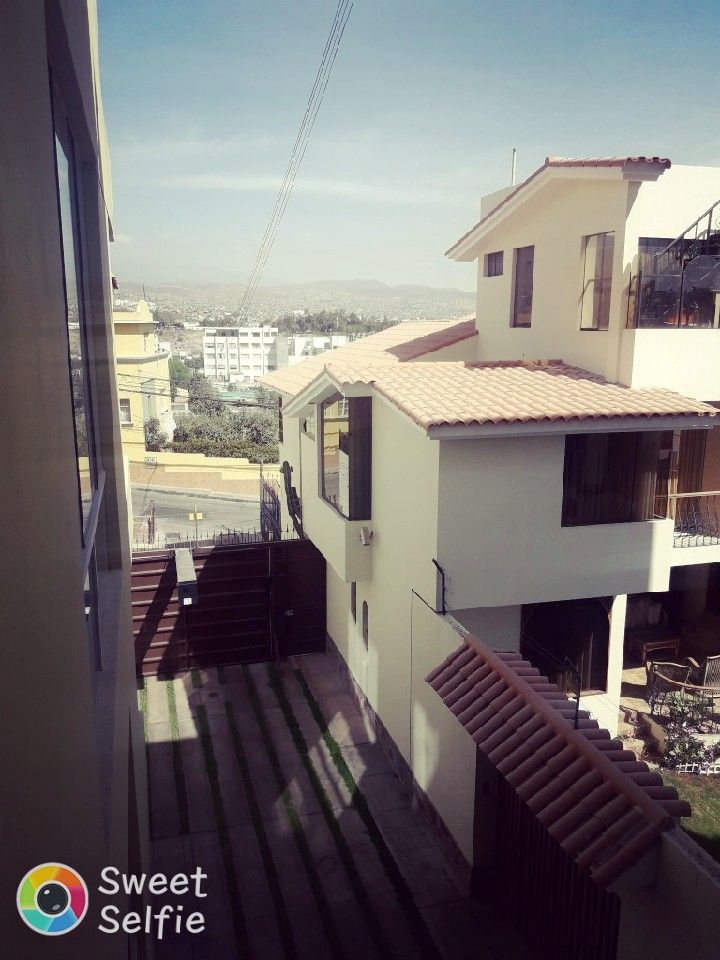 A view from the apartment reveals a grassy area bordered by modern white buildings. The terracotta tiled roof of the adjacent structure contrasts with the clear sky. Steps lead down to the green space, suggesting a well-maintained outdoor area.