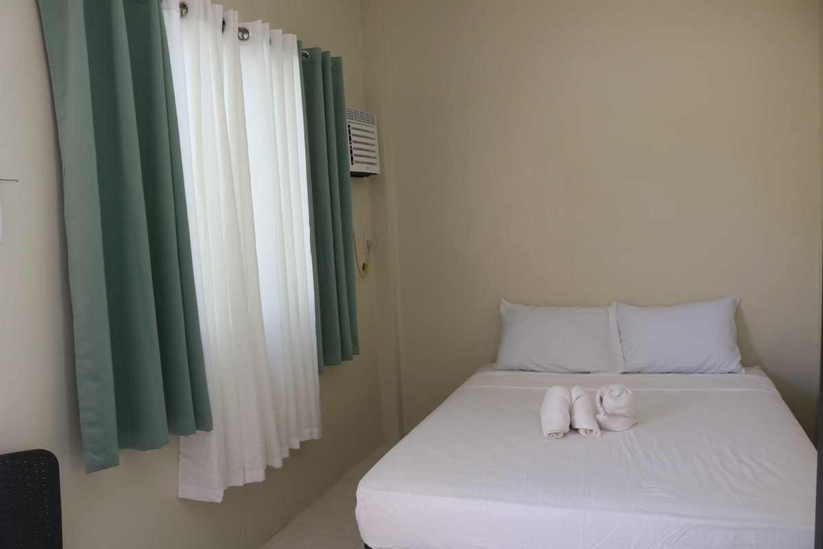 A well-lit room features a spacious bed with clean white linens. Two fluffy towels are neatly arranged at the foot of the bed. Natural light pours in through a window adorned with light green curtains, while an air conditioning unit is mounted on the wall.
