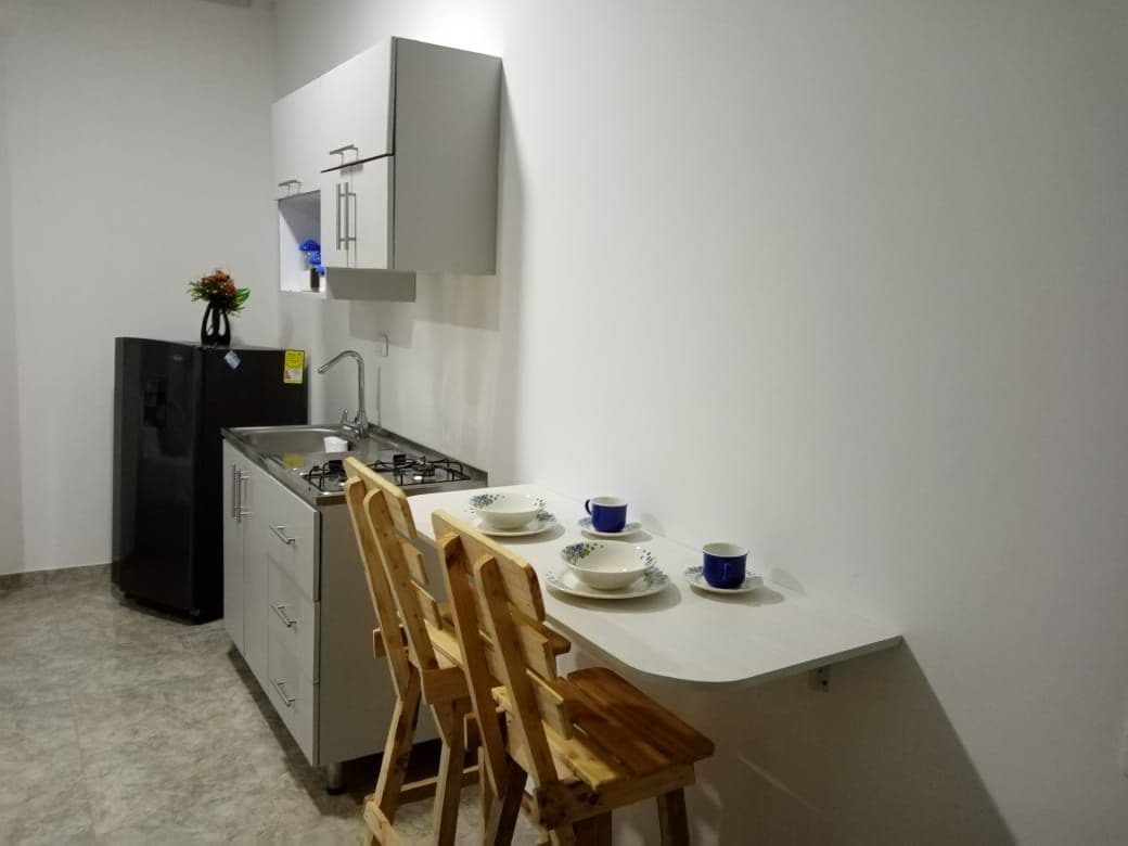 A modern kitchenette features sleek cabinetry and a gas stove. Two wooden stools are positioned at a small table set with blue and white dishware. A black refrigerator stands against the wall, and a small vase with flowers adds a touch of color.