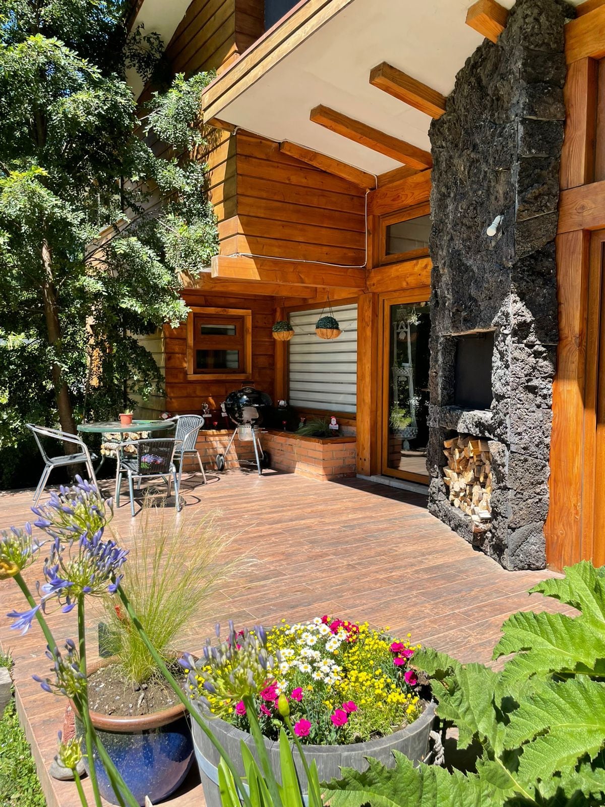 A wooden exterior is showcased, featuring a spacious patio area with a dining table and chairs. Potted flowers and greenery add color, while a stone fireplace is visible, enhancing the outdoor space's ambiance. Large trees provide shade, contributing to a tranquil environment.
