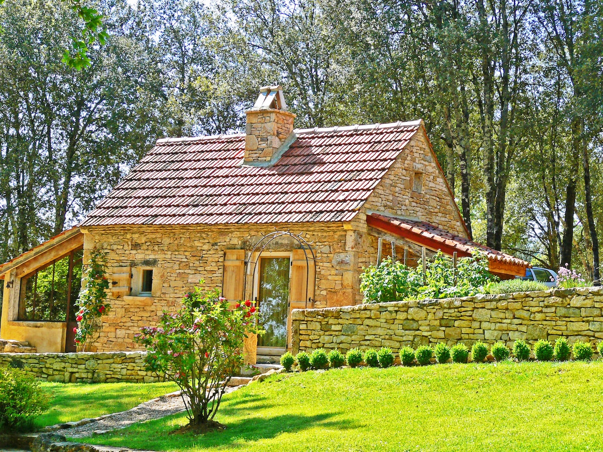 A charming stone cottage is surrounded by lush greenery, featuring a sloped roof with red tiles. The garden includes carefully trimmed hedges and colorful flower beds, enhancing the welcoming entrance. Large windows offer glimpses of the interior, contributing to the rustic appeal of the home.