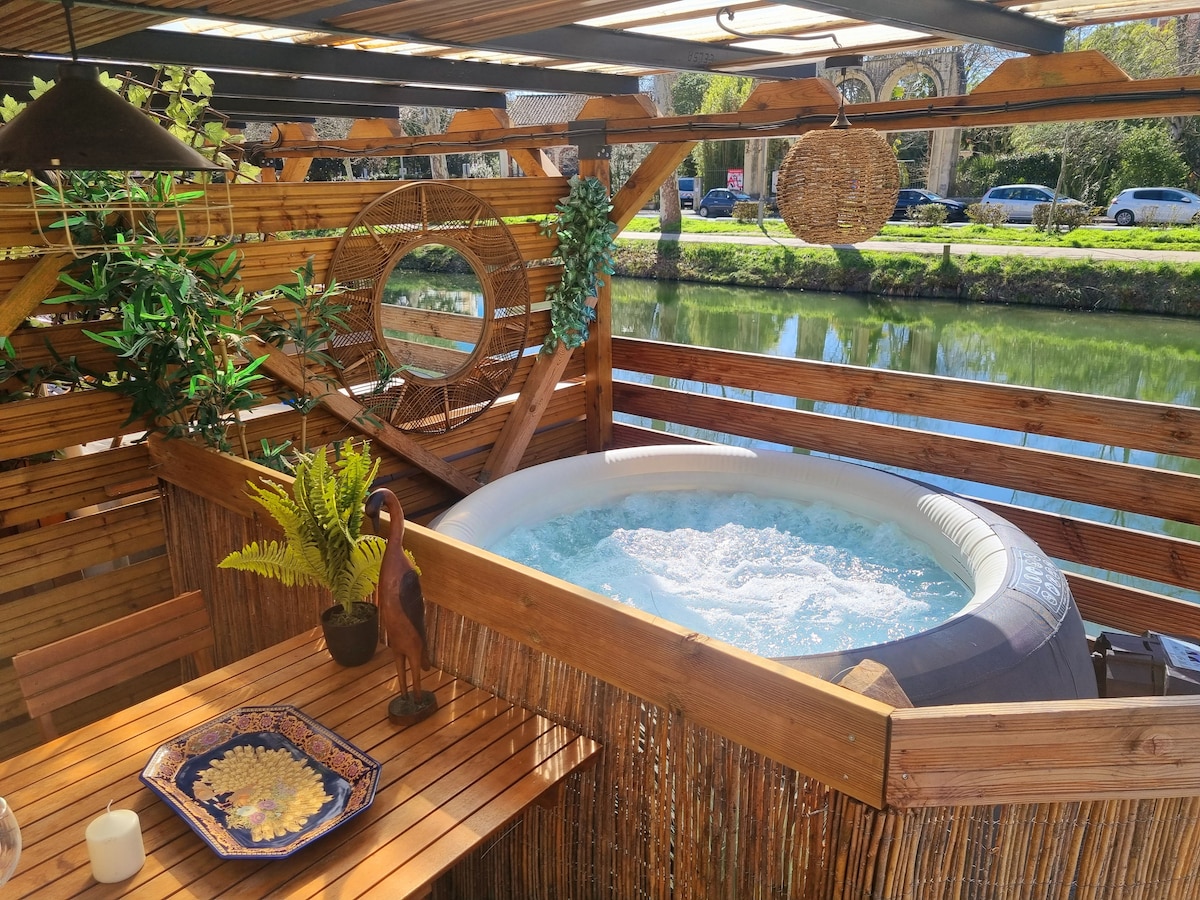 An inviting outdoor space features a private jacuzzi surrounded by wooden decking. Lush green plants are positioned nearby, and a dining table is set for enjoyment. The picturesque setting overlooks a serene canal, enhancing the tranquil atmosphere.