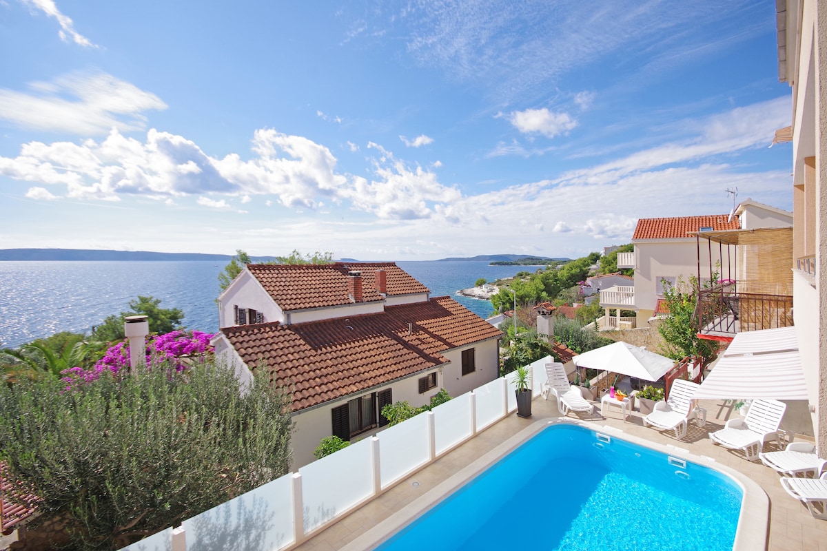 A serene view of the sea is captured from an elevated terrace. A clear blue pool is positioned in the foreground, surrounded by lounge chairs and shaded by umbrellas. Nearby, villas with traditional tiled roofs complement the lush greenery and vibrant flowers that adorn the landscape.