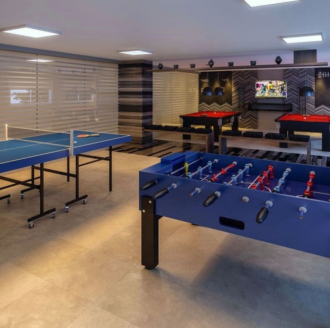 A game room features various entertainment options, including tables for ping pong and foosball, set against a backdrop of sleek, modern design. Soft lighting enhances the inviting atmosphere, with a large screen visible in the background for gaming or movies.
