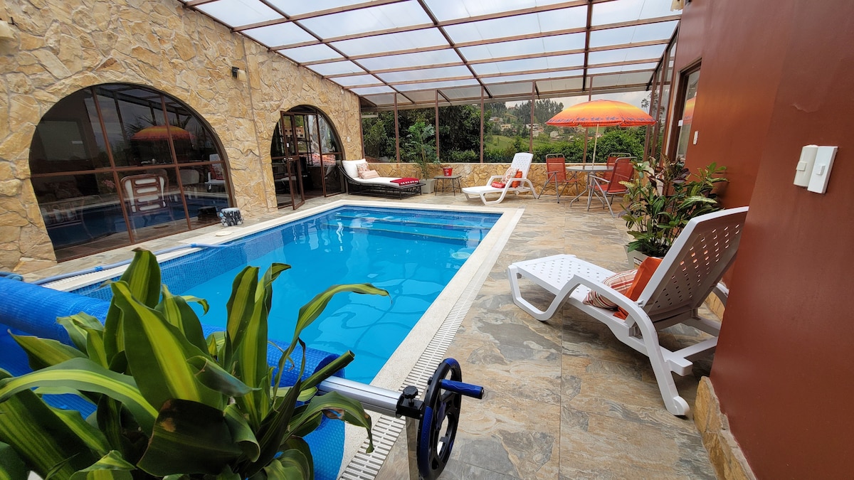 A covered swimming pool area is surrounded by stone walls and large arched windows. A few lounge chairs and a shaded seating area are visible, along with greenery placed around the pool. The space offers a blend of relaxation and nature.