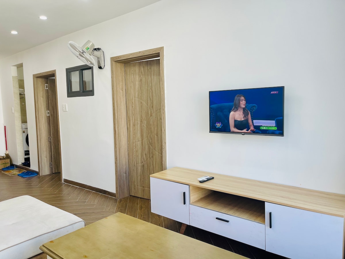 A comfortable living area is displayed with a light wood TV stand and a wall-mounted television. Two doors lead to other rooms, framed by neutral walls. Ambient lighting enhances the clean design, while a small remote control is visible on the TV stand.