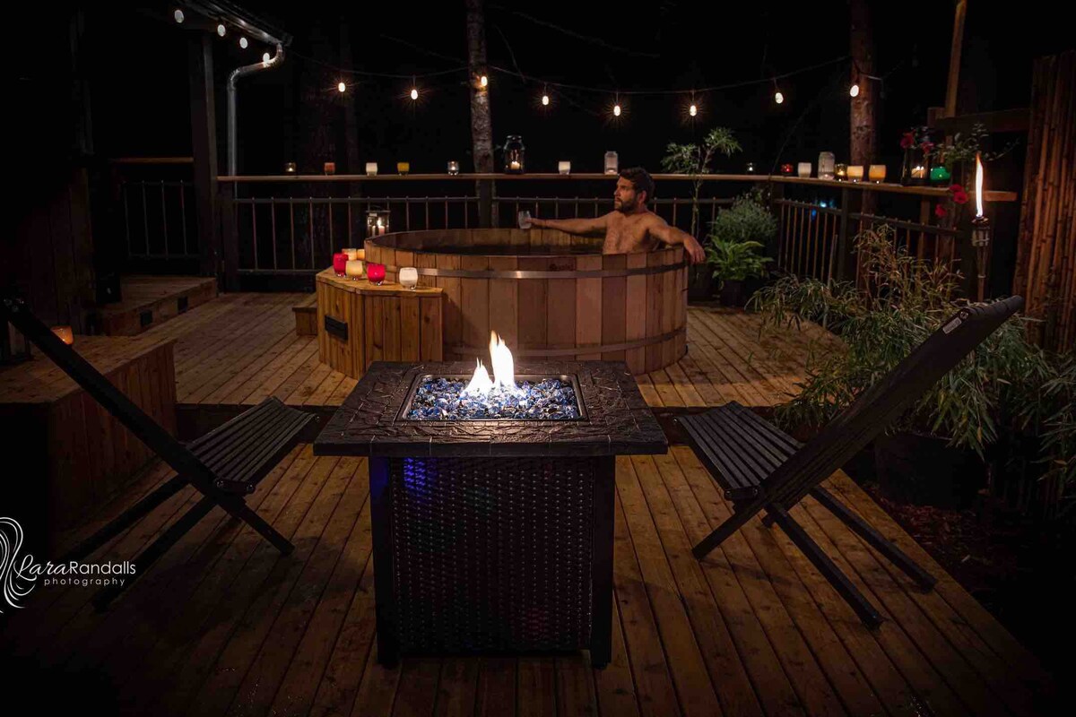 A wooden deck features a circular hot tub surrounded by warm lighting and decorative candles. A smokeless fire pit with glass stones is centrally placed, accompanied by two lawn chairs. Lush greenery is visible in the background, enhancing the tranquil ambiance.