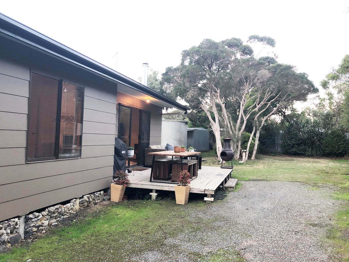 The exterior of the home features a wooden deck with a dining table and chairs, accompanied by a chiminea and a barbecue grill. Surrounding greenery complements the serene outdoor space, while large windows on the house provide natural light to the interior.