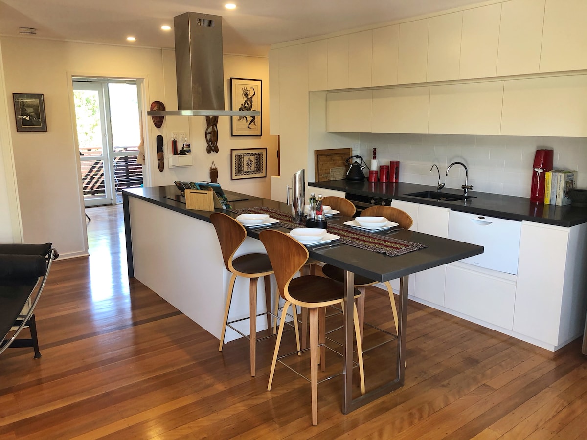 A contemporary kitchen features sleek cabinetry and a spacious island with bar stools. Natural light brightens the area through nearby windows. The layout includes modern appliances and an inviting seating arrangement, creating a functional yet stylish space for dining and socializing.