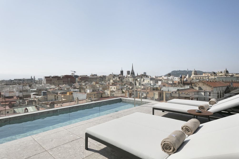A spacious rooftop terrace presents sleek loungers with rolled towels, overlooking the diverse skyline of the city. The horizon features historic spires and modern buildings, while a shallow pool reflects the sky, creating a serene spot for relaxation and enjoyment.