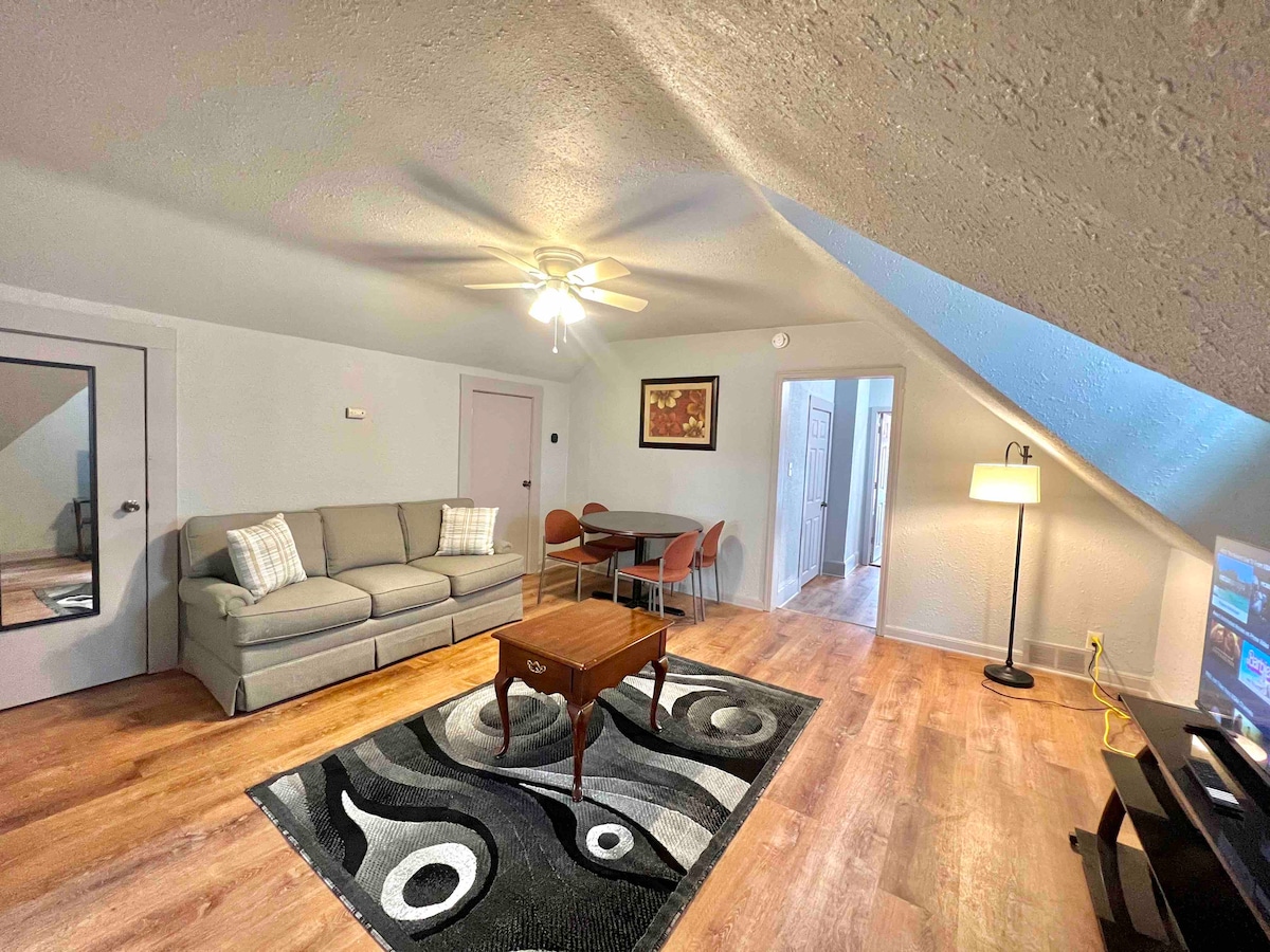 A living space features a light-colored sofa, a coffee table, and a dining area with four chairs. Natural light enters through a skylight, illuminating a patterned area rug. A ceiling fan is present, and a television is positioned against the wall.