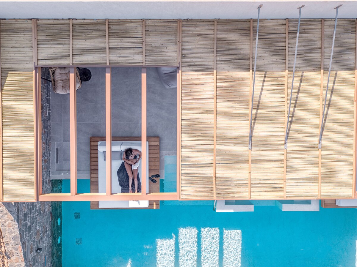 An aerial view highlights the private infinity pool surrounded by a wooden terrace. A daybed is positioned nearby, providing a comfortable spot for relaxation. The pool’s turquoise water reflects the sunlight, enhancing the serene ambiance of the space.