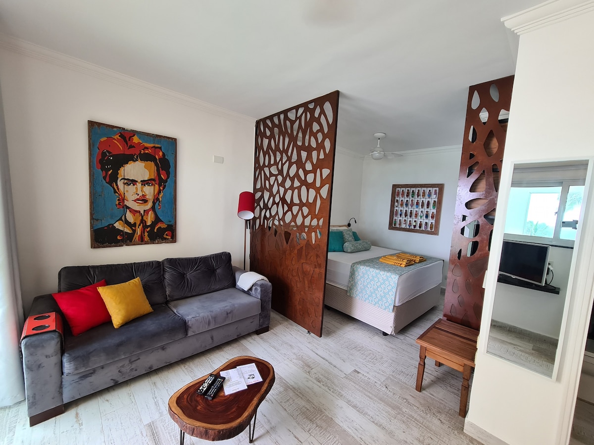 A modern living area is visible, featuring a dark sofa adorned with a colorful throw pillow. A wooden partition divides the space, revealing a light-colored bed with layered bedding. Artwork with vibrant colors and a lamp are included in the decor, creating a welcoming ambiance.