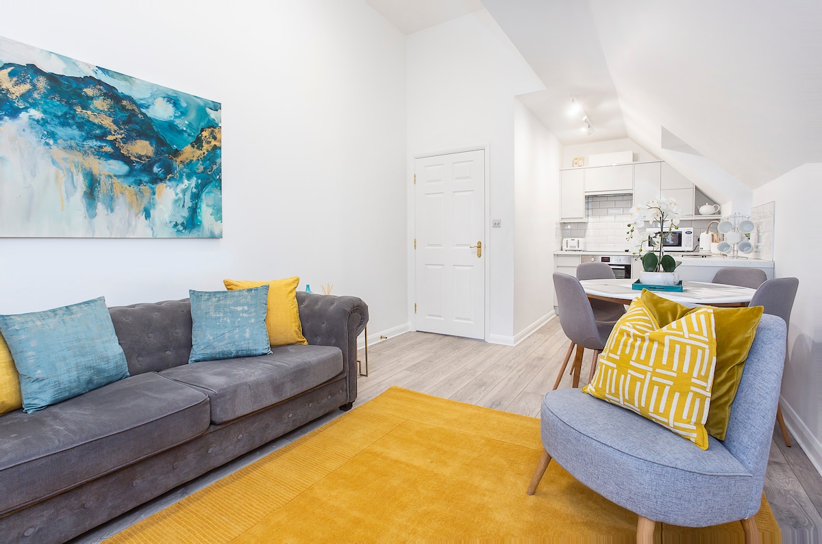 The living area features a modern gray sofa adorned with colorful cushions in shades of blue and yellow. A bright yellow rug complements the space, while a large abstract painting adds a focal point on the wall. The dining area is visible in the background, showcasing a modern table and chairs.