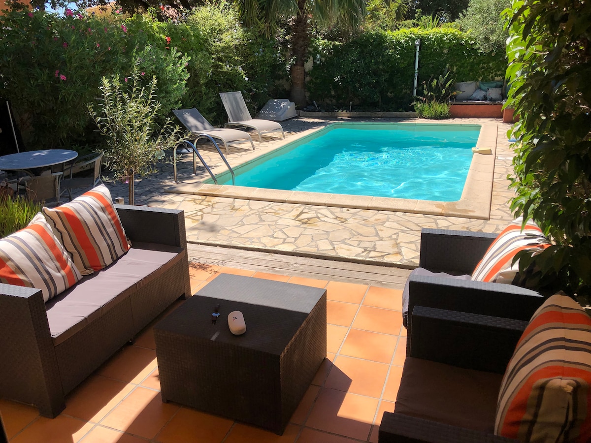 Seating is arranged around a low table on a sunlit terrace, providing a view of the private swimming pool. Lounge chairs are positioned alongside the pool, surrounded by lush greenery and flowers, creating a serene outdoor relaxation area.