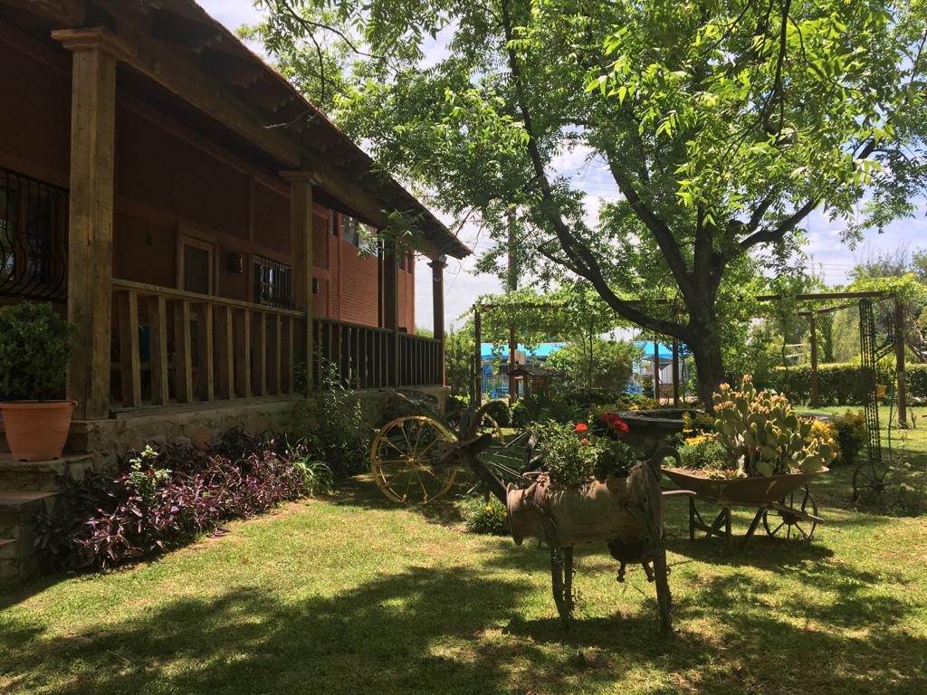 A rustic cabin is nestled beneath lush trees, surrounded by a graceful garden. Decorative elements include an old cart and potted plants, while vibrant green grass enhances the outdoor area. The inviting porch extends from the cabin, offering a peaceful view of the serene surroundings.