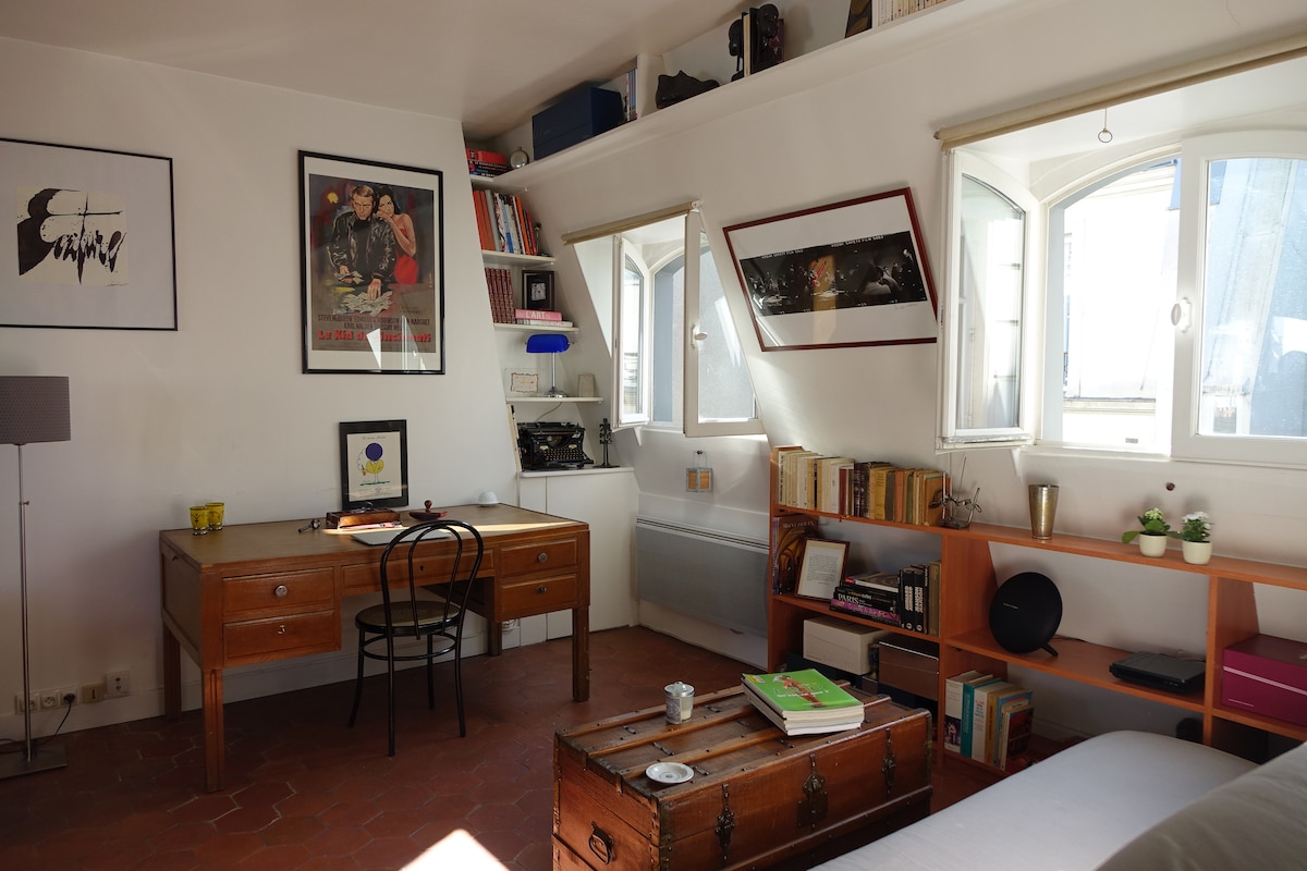 A bright, airy room features a wooden desk with a black chair positioned near a window. Shelves filled with books are seen along one wall, while framed artwork adds character. Light enters through multiple windows, enhancing the inviting ambiance of the space.