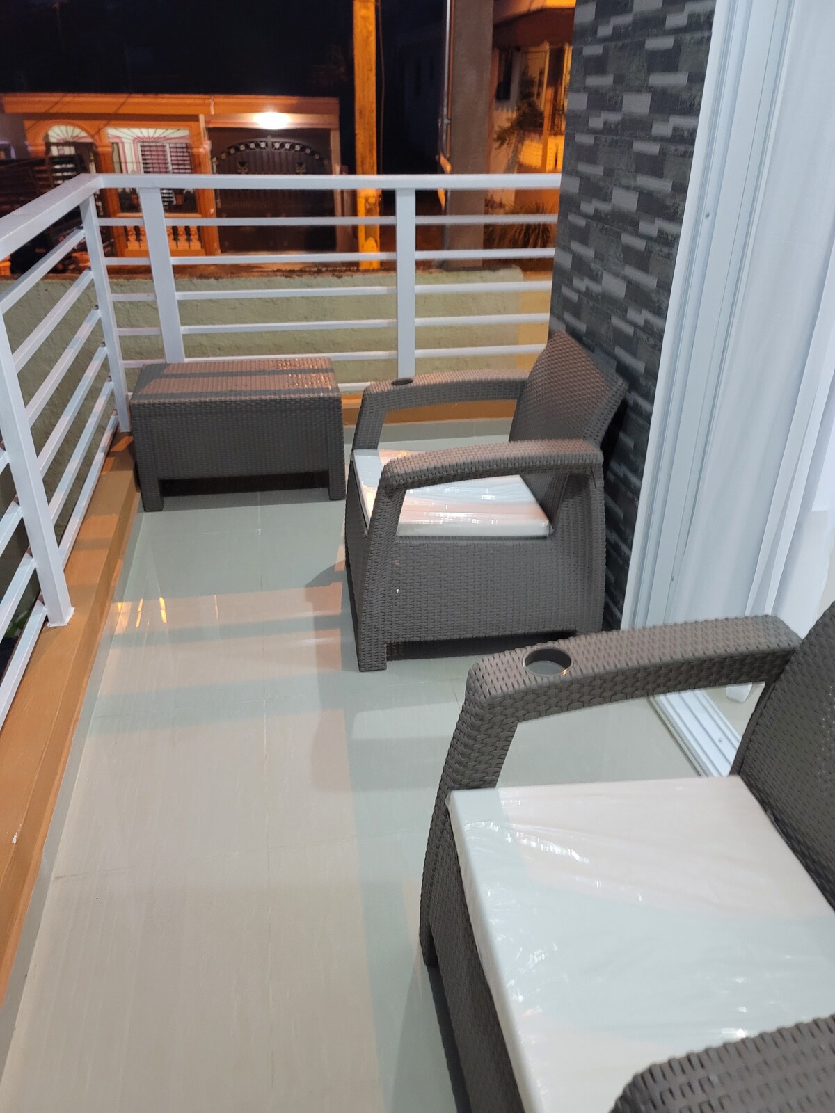 A balcony area is furnished with two rattan-style chairs and a small table. The sleek flooring enhances the outdoor space, while a railing provides security. Soft lighting from nearby structures adds a cozy ambiance during the night.