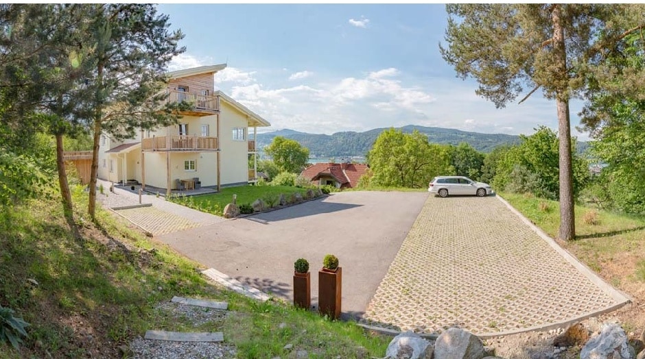An exterior view of the villa reveals a spacious parking area bordered by greenery. The building, featuring multiple balconies, is set against a backdrop of rolling hills and trees, with scenic valley views under a clear sky.