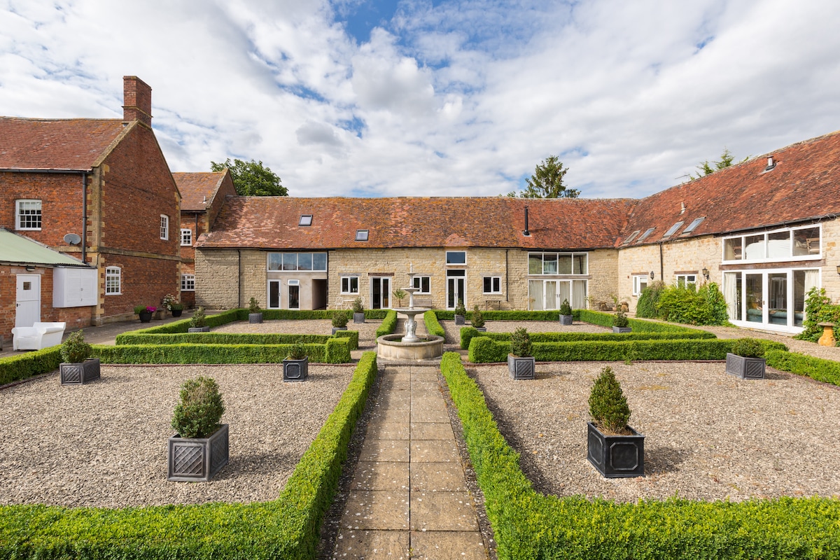 A tranquil courtyard is presented, featuring well-maintained hedges and gravel pathways. A central fountain stands surrounded by neatly arranged planters. The building's facade, composed of stone and brick, offers a blend of traditional and contemporary design, with large windows allowing natural light to enter.