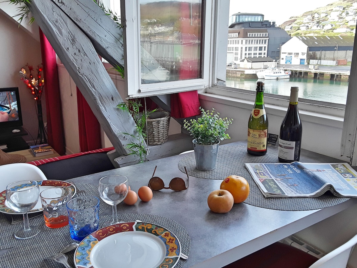 A dining table is set for a meal, featuring plates and glassware. Two bottles of wine are placed alongside fruits. A newspaper lies on the table, with a view of the harbor visible through an open window framed by wooden beams.