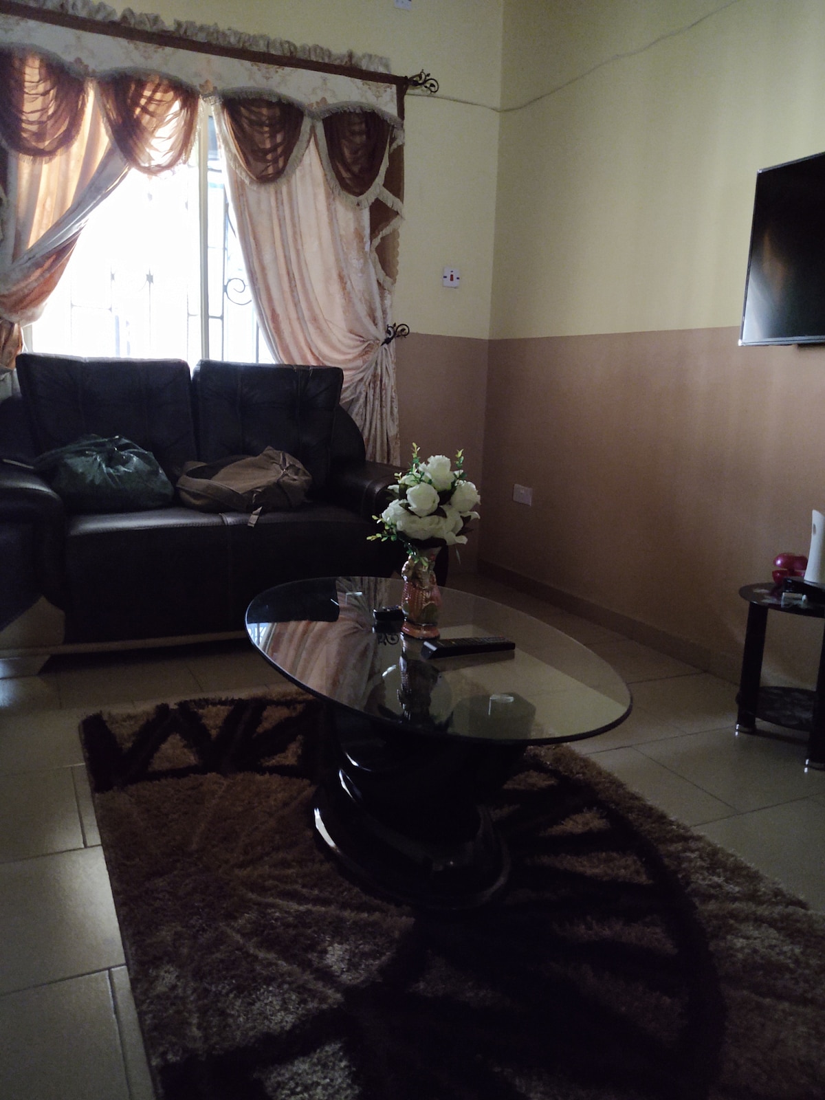 The living room features a dark leather sofa positioned beside a light-colored wall. A glass-top coffee table is centered on a patterned rug, while a decorative vase with flowers adds a touch of elegance. Natural light filters through sheer drapes, illuminating the space.