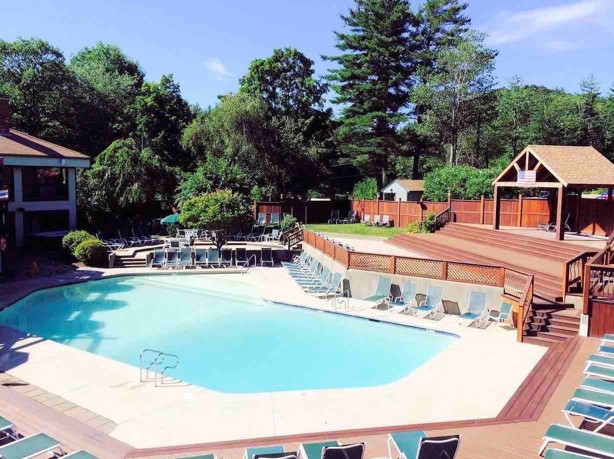A spacious outdoor pool area is framed by an expansive wooden deck and surrounded by lounge chairs. Lush greenery is present in the background, while a shaded gazebo offers an inviting space for relaxation.
