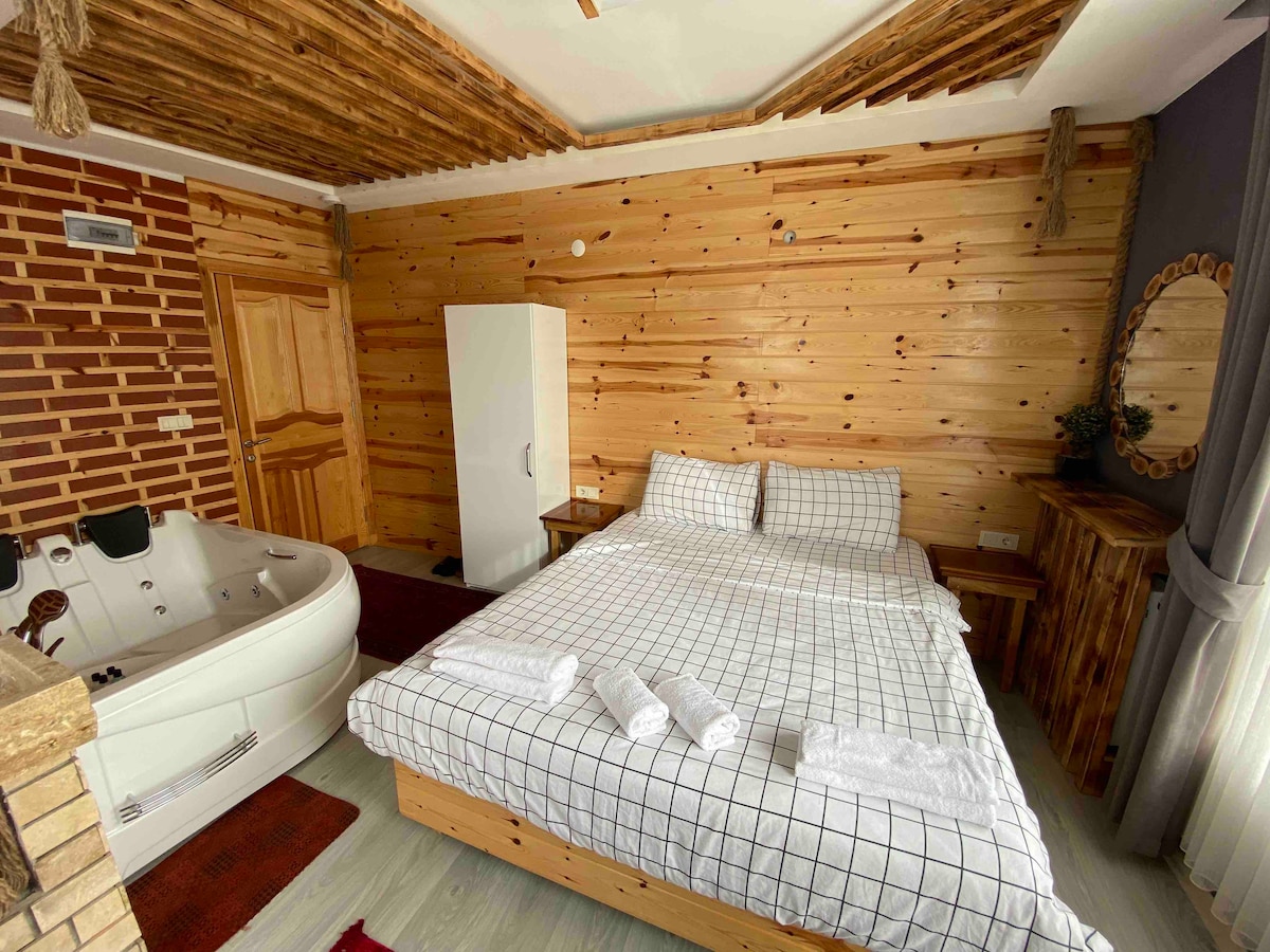 A warm, wooden interior is featured, with a spacious bed dressed in a checkered pattern. A whirlpool tub is positioned nearby, alongside a white wardrobe. Natural light enters through the window, illuminating the textured walls made of wood.