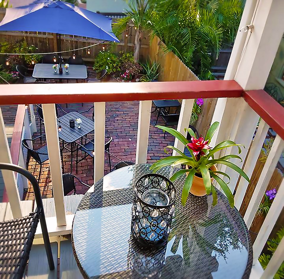 An outdoor seating area is visible, featuring a round table adorned with a decorative black candle holder and a potted plant with vibrant flowers. The brick courtyard below is surrounded by greenery and twinkling string lights, creating a peaceful atmosphere.