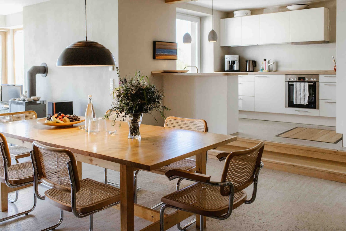 An open-plan kitchen and dining area is showcased, featuring a wooden dining table surrounded by several chairs. A modern pendant light hangs above the table. The kitchen is equipped with white cabinetry, appliances, and a large window allowing natural light to illuminate the space.