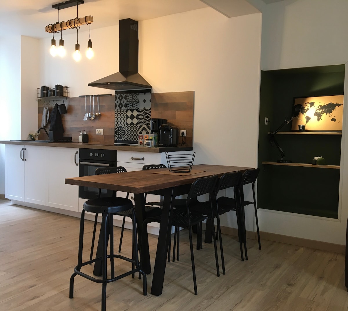 An open kitchen area features a wooden dining table surrounded by black metal chairs. A stylish backsplash with patterned tiles complements modern appliances, including a stovetop and coffee maker. A small nook with a wall-mounted lamp and a world map adds a touch of design.