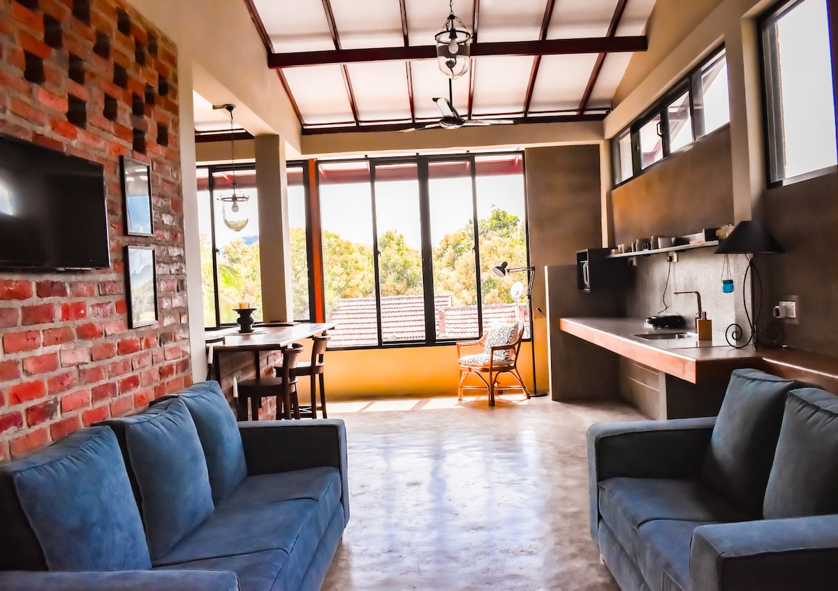 An open-concept living area features two plush sofas arranged for socializing. Large windows allow natural light to illuminate the space, showcasing views of lush greenery. A dining table and a compact kitchen are visible, creating a functional and inviting environment.
