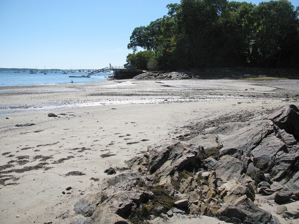 A sandy beach stretches along the coastline, with rocky outcrops visible on the left. Calm water reflects the blue sky, dotted with moored boats in the distance. Lush greenery borders the shoreline, providing a peaceful backdrop for outdoor activities.
