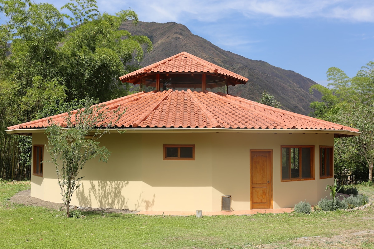 The round structure features a terracotta tiled roof and large windows, allowing natural light to fill the space. Surrounded by lush greenery, the house is set against a backdrop of mountains, highlighting its tranquil location. An inviting wooden door is centrally positioned at the front.