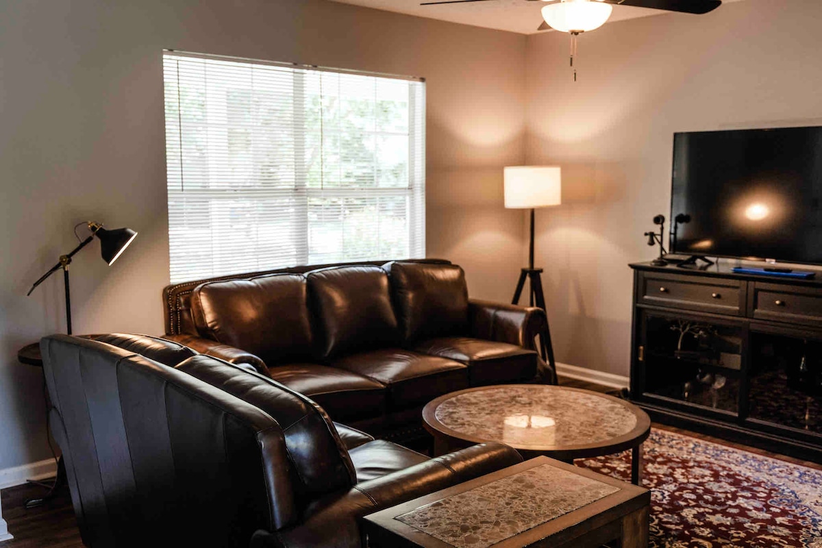 A well-lit living area features a dark leather sofa and a pair of armchairs, arranged around a round table. A floor lamp and a television stand are also present, with a large window providing natural light and a view of the outside.