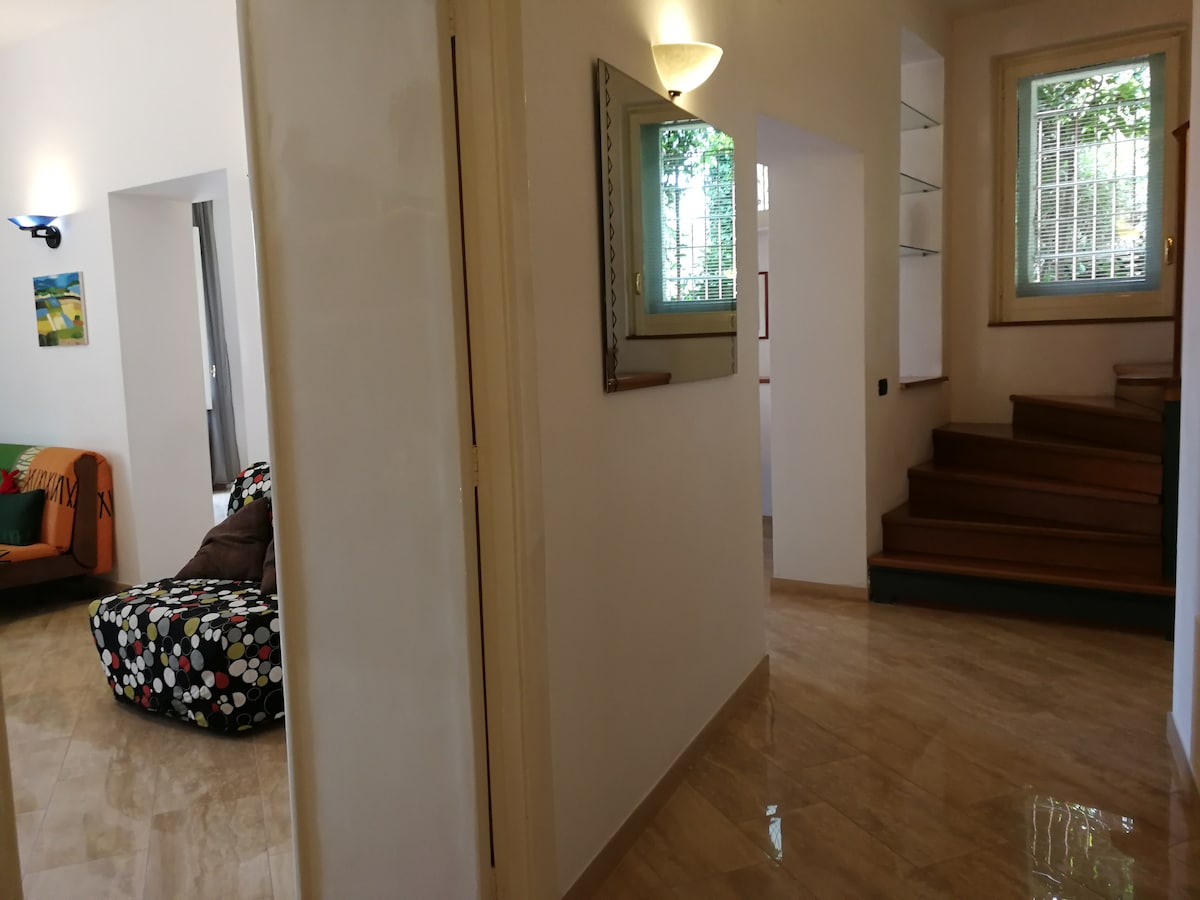 A bright hallway leads to various spaces within the apartment. To the left, a lounge area features a colorful patterned bean bag and a framed artwork. A wooden staircase ascends to the upper level, while natural light pours in through the windows.