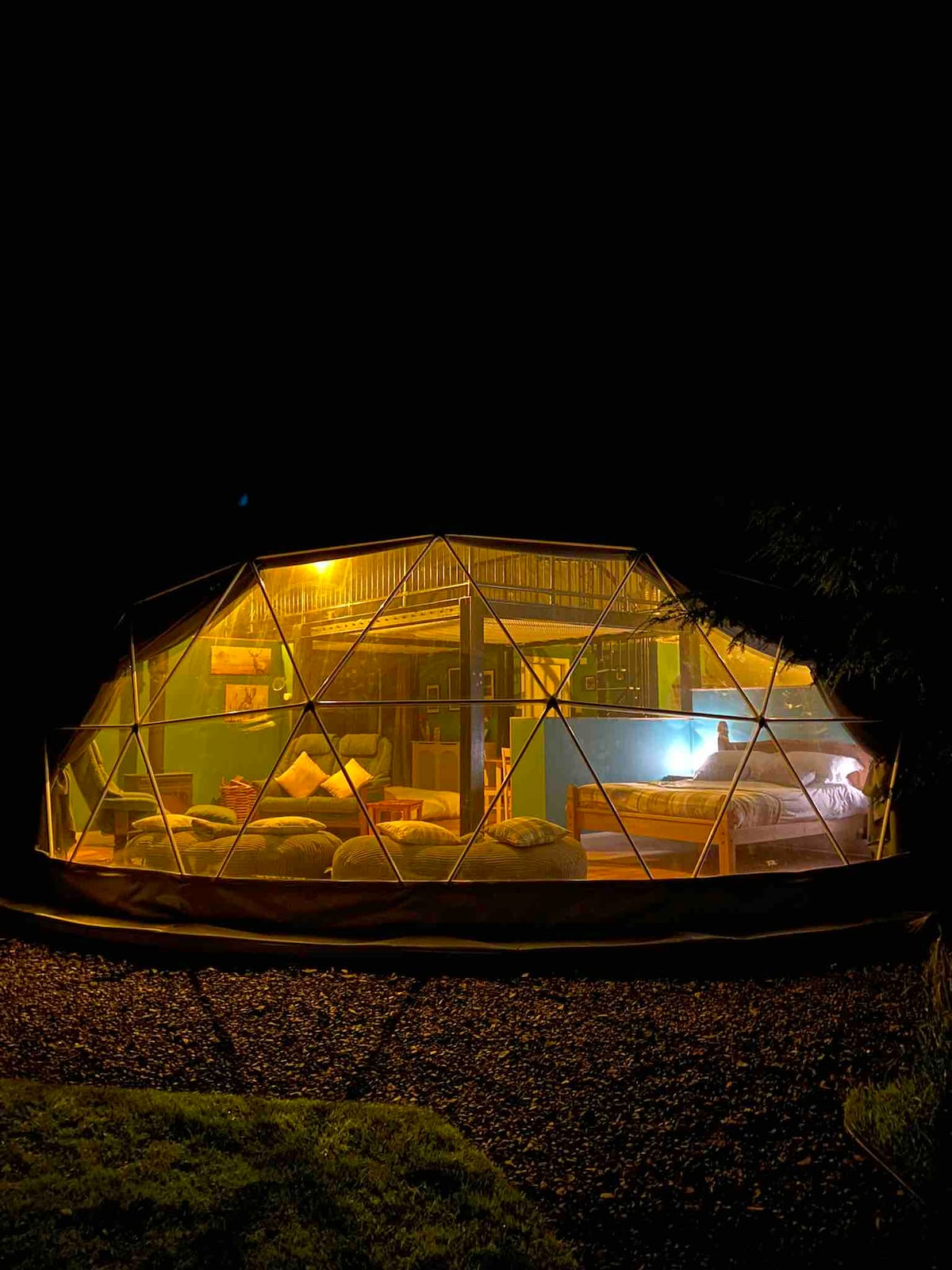 The geodesic dome is illuminated from within, showcasing a cozy living area with soft seating and a spacious bed. Large windows provide views of the surrounding forest while creating a connection to the natural environment. The warm light enhances the welcoming atmosphere at night.