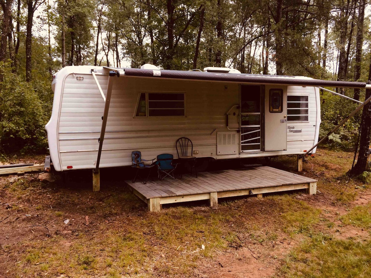 A travel trailer is situated on a wooden deck, surrounded by dense trees. The exterior is white, featuring a small entry step and an awning providing shade. A chair is placed in front, contributing to the rustic setting of the private woodland.