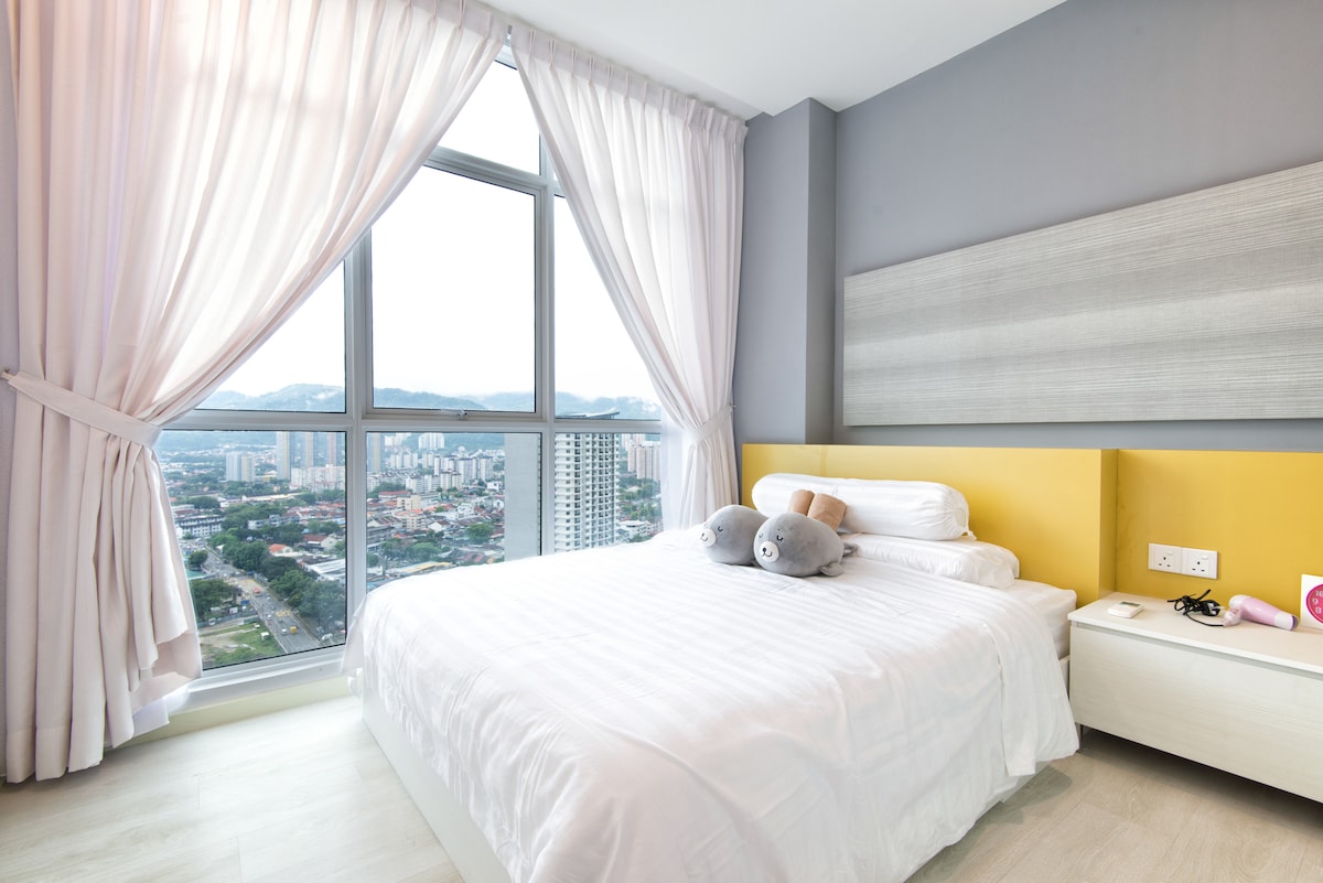 A well-lit bedroom features a spacious bed adorned with two plush toys. Sheer curtains frame a large window, allowing for scenic views of the cityscape. The soft color scheme incorporates calming tones, while a minimalist nightstand holds a few personal items.