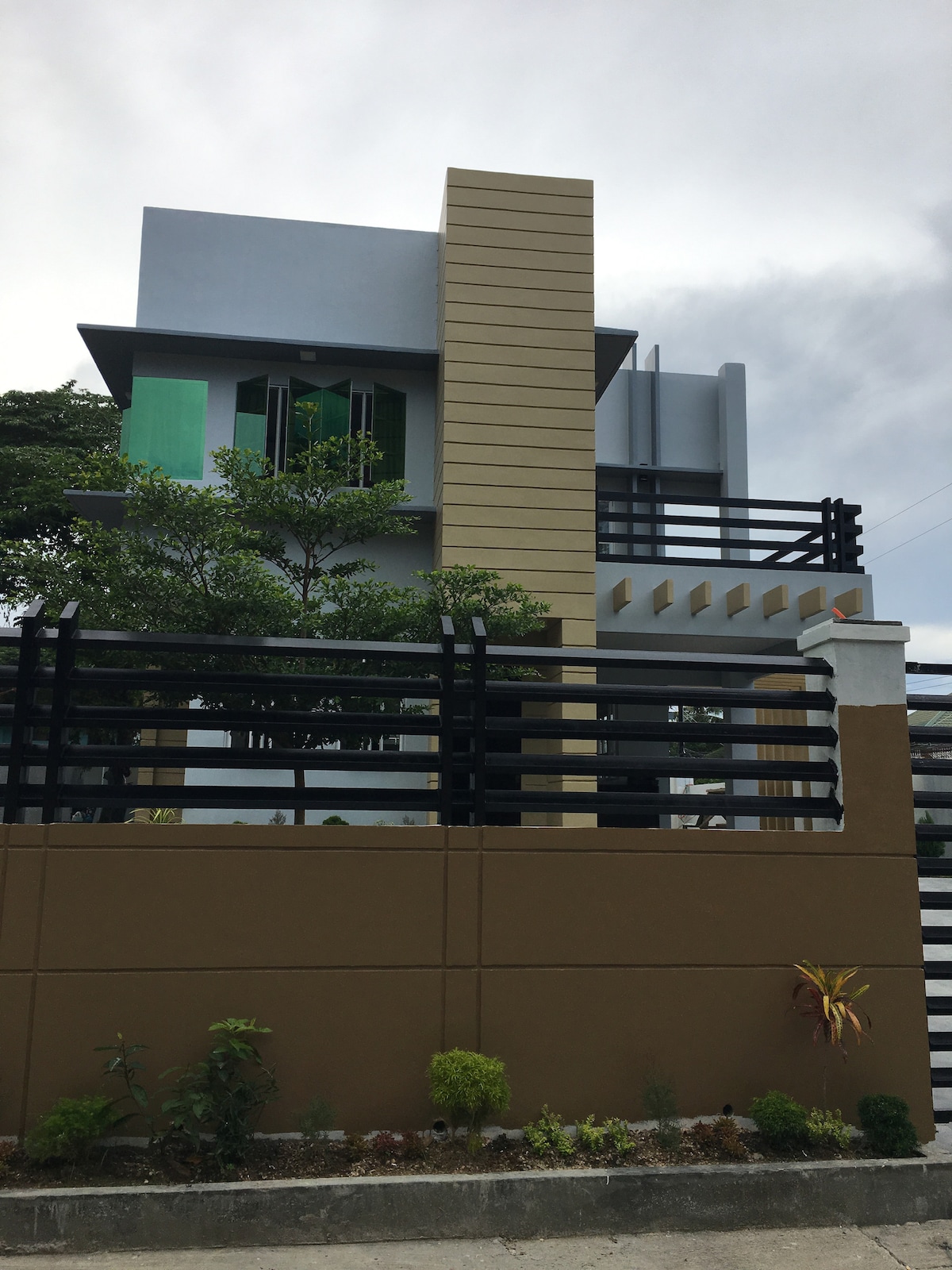 A modern two-story house is visible, featuring a mix of bold colors and textures. Large windows reflect natural light, while green accents on the second floor add a touch of warmth. A structured fence surrounds the property, emphasizing a sense of privacy and security.